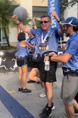 Dwayne crossing the finish line of a 5K, race number 255, both arms raised, with Chaim Backman beside him