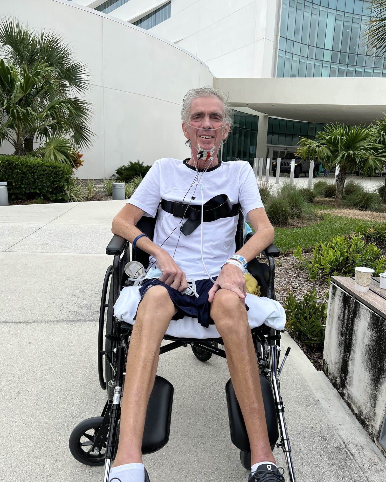 Dwayne outside the hospital in a wheelchair, on oxygen through a nasal cannula, smiling at the camera