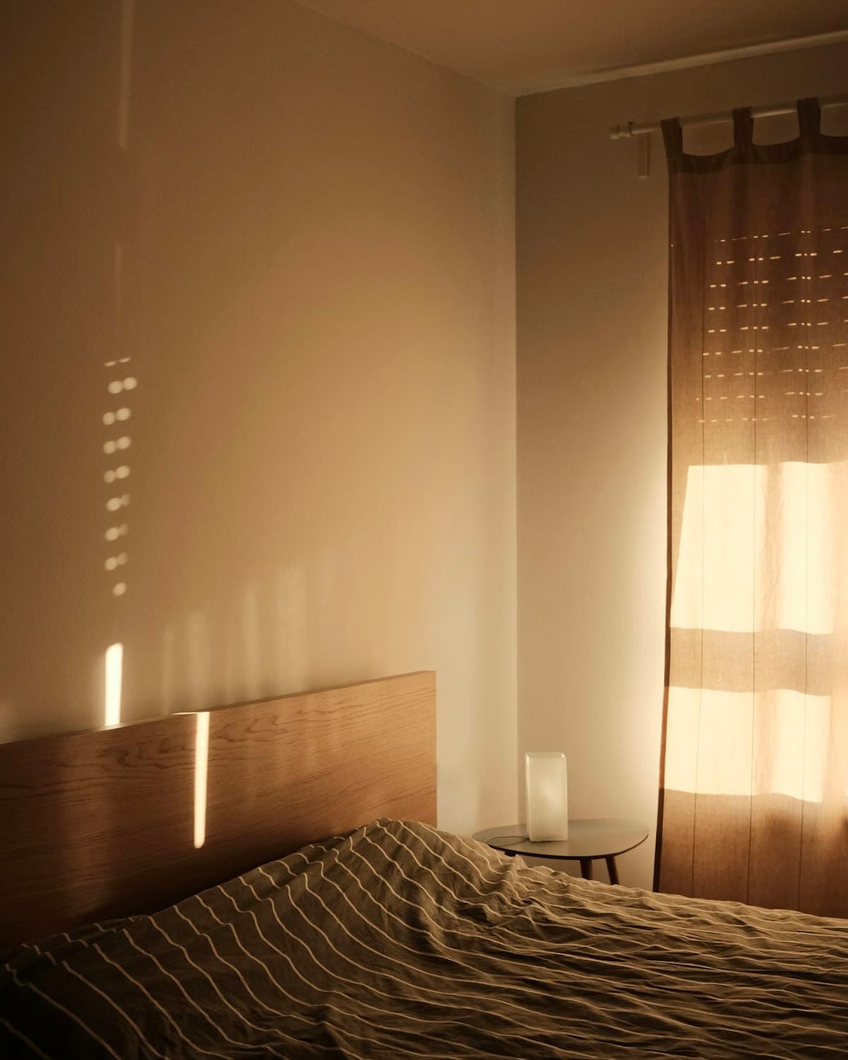 Morning light falling across a made bed and a small bedside lamp