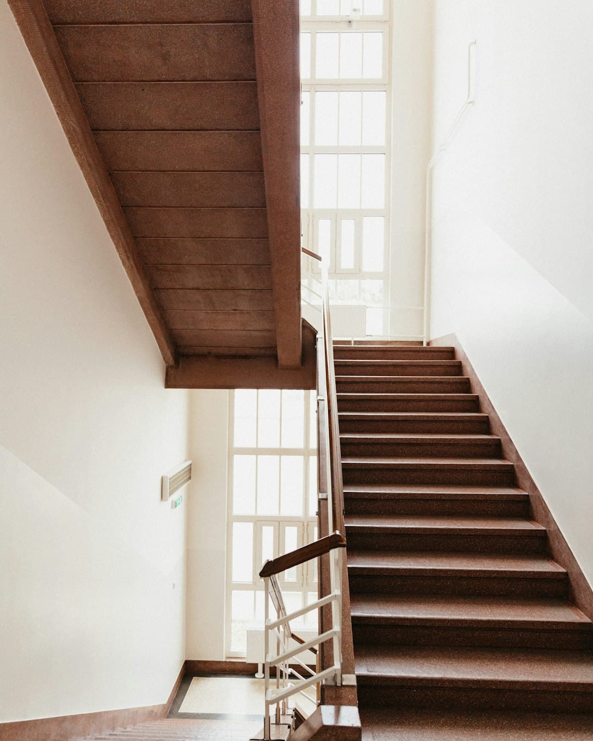 A stairway with a wooden handrail, climbing past tall sunlit windows